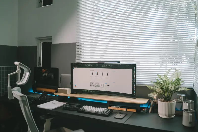 A modern office desk setup featuring a computer monitor, ergonomic chair, and potted plant by a window
