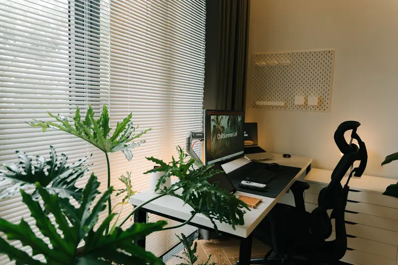A stylish home office with a desk, chair, and lush green plants near a window with blinds