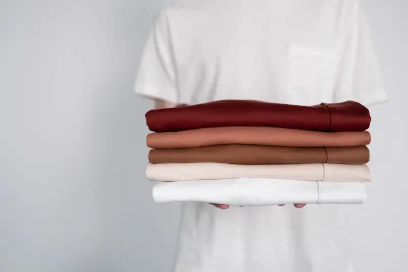 A person holding a stack of neatly folded shirts in earthy tones against a soft white background